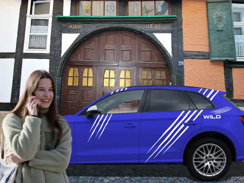 Glückliche junge Frau telefoniert vor ihrem blauen Audi Q5 mit Sticker-Motiv Krallen Kratzspur in Weiß, im Hintergrund altes Fachwerkhaus