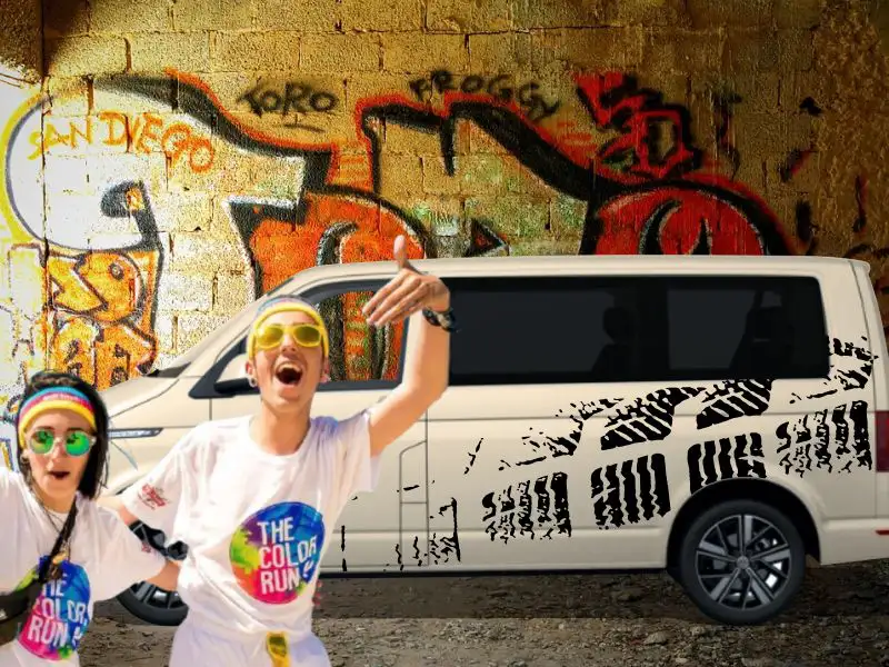 Fröhliches junges Paar mit bunten Sonnenbrillen vor einem Kleinbus VW T6 Bulli mit dem Sticker-Motiv Tire Track Reifenspur in Schwarz, der vor einer bunten Graffiti-Hauswand steht